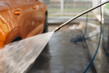 Driver using pressure washer to clean orange car at a wash bay during a sunny day