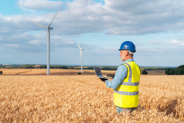 Energy engineer in safety gear uses laptop in field near wind turbines, showcasing wind energy green technology