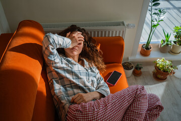 Naklejka na ściany i meble Woman feeling unwell and exhausted while lying on an orange sofa, covering her eyes with a hand, suffering from toxicosis, nausea, or a headache, indicating discomfort and illness Naklejka na ściany i meble Woman feeling unwell and exhausted while lying on an orange sofa, covering her eyes with a hand, suffering from toxicosis, nausea, or a headache, indicating discomfort and illness