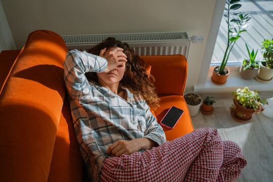 Fototapeta Woman feeling unwell and exhausted while lying on an orange sofa, covering her eyes with a hand, suffering from toxicosis, nausea, or a headache, indicating discomfort and illness