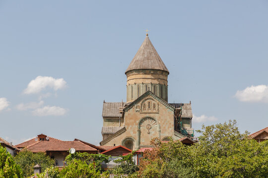 View of an ancient stone cathedral rises above the rooftops and lush greenery, under a soft blue sky, in Mtskheta, Mtskheta-Mtianeti, Georgia.