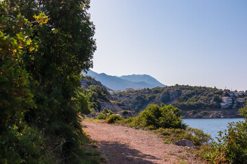 A rustic dirt path meanders through lush green Mediterranean foliage and trees, leading towards coastal village and the majestic Velebit mountains in the background, with the blue sea peeking through