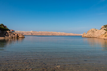 Fototapeta premium Crystal-clear turquoise water in a serene Croatian bay near Karlobag reveals a pebbled seabed, with rocky shores and the distant silhouette of Pag Island creating a peaceful coastal scene.