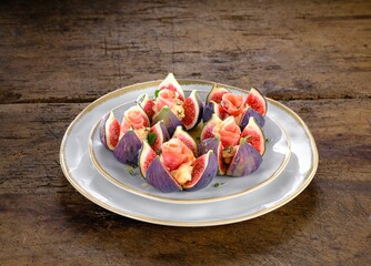 Ripe dark figs are cut into quarters and topped with soft cheese. Delicate prosciutto roses are placed on top, arranged on a decorative plate on a textured wooden table