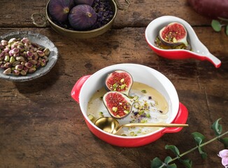 Several dishes are arranged on a rustic wooden table. One bowl contains figs in a sweet soup, another holds pistachios, and there is a small dish with a single fig