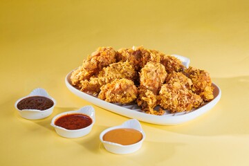 A platter piled high with crispy, golden fried chicken wings is displayed alongside three small dishes filled with tempting dipping sauces against a sunny yellow background