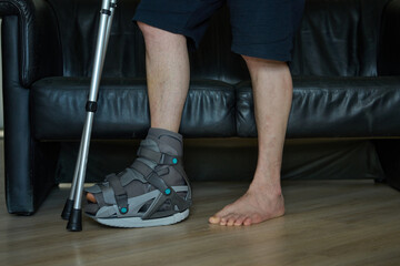 Person with injured leg standing with crutch indoors. Medical boot, crutch, weight on healthy leg. Recovery after injury.