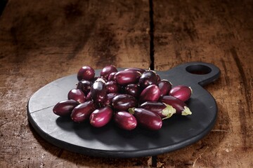 Small, shiny eggplants are piled high on a black, round serving board. The board rests on a weathered wooden surface, showcasing the fresh produce