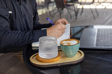 A person is sitting at a table with a laptop and two drinks, writing something.