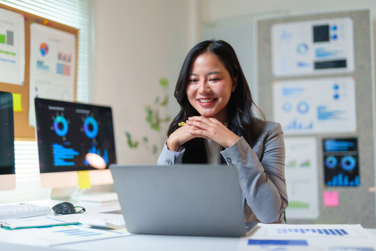 Asian businesswoman smiling, connecting with colleagues online, reviewing business reports and charts on laptop and monitor screens