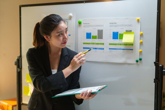 Young businesswoman pointing at financial charts on whiteboard, presenting data during a business meeting in a professional office
