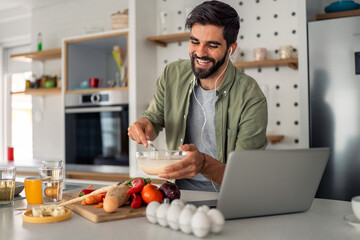 Smiling man mixing ingredients in modern kitchen with laptop fresh vegetables and coffee cup