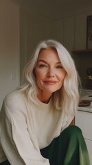A woman with white hair is sitting in a kitchen. She is smiling and looking at the camera