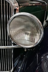 Detail off round vintage headlight in chrome frame on oldtimer Czechoslovak limousine from year 1930. Front mask chrome grille in background. 