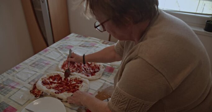 Elderly woman spreading tomato sauce on pizza dough. Grandma making pizza for her family. A senior woman cooking in her kitchen.