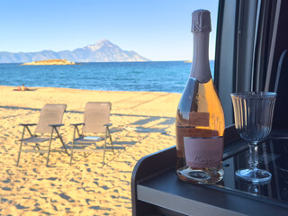 A bottle of chilled rosé wine and a glass by a camper van overlooking a golden sandy beach and Mount Athos on Halkidiki, Greece. Relaxed coastal travel mood with soft pastel light.