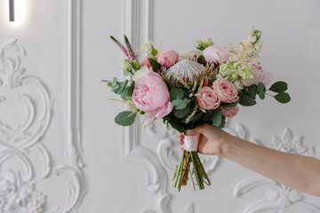Hand holding a bold, modern bridal bouquet with pink peonies, blush roses, King Protea, and eucalyptus against an ornate classic white stucco wall