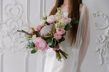 Elegant bride in a white satin gown holding a bold mixed bouquet of pink peonies, blush roses, and a large gray-pink King Protea against a classic white stucco wall
