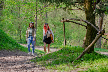 Two happy young women enjoying a leisurely walk on a forest path, surrounded by vibrant green nature. Friendship and outdoor recreation on a sunny day.