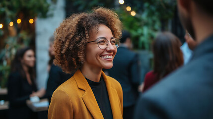 A businesswoman with curly hair and glasses smiles at a coworker at an office meeting.