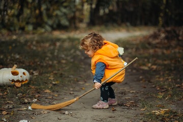 A young child in a pumpkin costume sweeps with a broom on a forest path during Halloween, with a carved pumpkin nearby