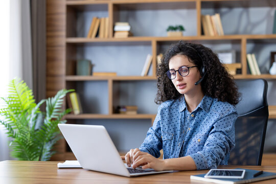 Young woman with curly hair wearing a headset with a microphone. Concentrating while typing on a laptop. Working remotely from her modern home office setup with a bookshelf and plant in the background
