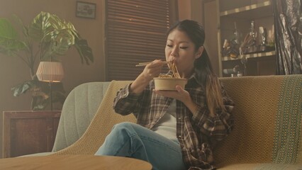 A Korean woman is sitting comfortably on a sofa, using chopsticks to eat Asian noodles from a bowl.