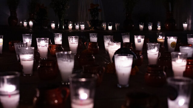 Gran conjunto de veladoras tradicionales encendidas en vasos de vidrio y jarritos de barro en la oscuridad para ofrenda