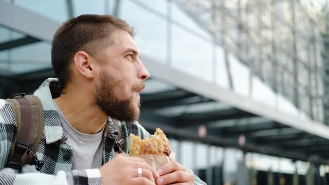 Young man with a beard and backpack enjoys eating traditional Serbian burek outdoors near a modern glass building