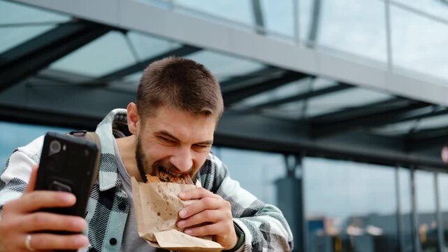 Young man with a beard and backpack eats traditional Serbian burek while talking on a video call outdoors near a modern glass building