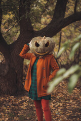 A woman in an orange coat and plaid dress with a carved pumpkin mask poses in an autumn forest on Halloween.