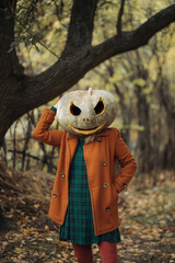 A woman in an orange coat and plaid dress with a carved pumpkin mask poses in an autumn forest on Halloween.