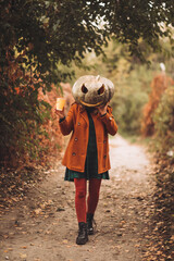 A woman in an orange coat and plaid dress with a carved pumpkin mask poses in an autumn forest on Halloween.