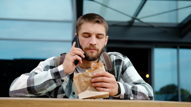 Young man with a beard and backpack eats Serbian burek while talking on his smartphone outdoors near a modern glass building