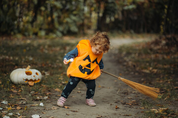 A young child in a pumpkin costume sweeps with a broom on a forest path during Halloween, with a carved pumpkin nearby