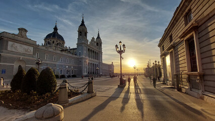 People stroll along the plaza near Almudena Cathedral in Madrid at sunset, casting long shadows on...