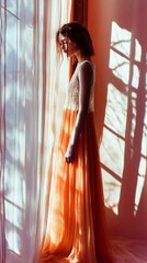 A woman in a long orange dress is standing in front of a window. The image has a warm and inviting mood, with the orange dress