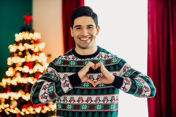 Smiling young man wearing a festive sweater making a hand heart gesture in front of a beautifully decorated Christmas tree