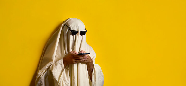 Halloween. Ghosting. Ghost costume with white blanket and sunglasses texting on smartphone over yellow background