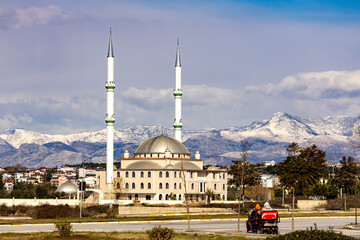 White mosque with twin minarets stands against snow-dusted Taurus Mountains under a clear winter sky, showcasing winter serenity in Antalya. Ilica, Turkey.

