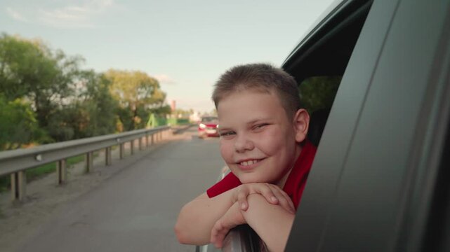 Child smiles and rejoices in family vacation, car trip. Little boy is sitting in car, look from an open window, smile of child. Happy boy rides in car, looks out of window, enjoys landscape, warm wind