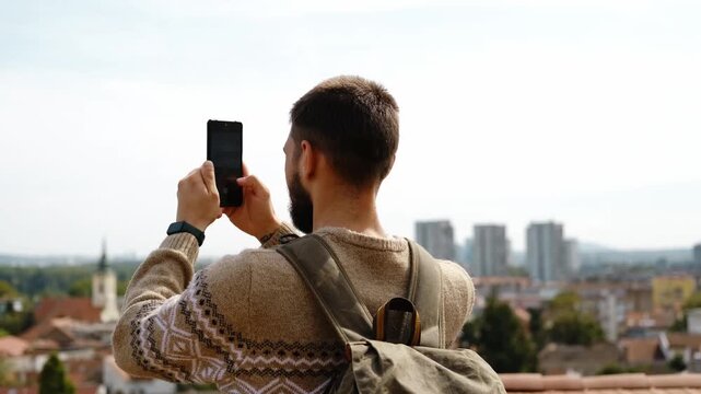 A man with a backpack takes pictures or making video of Zemun rooftops and the Danube River using his smartphone on a sunny day. Travel blogger concept