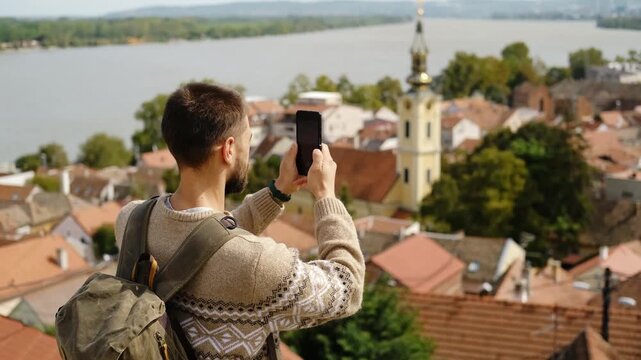 A man with a backpack takes pictures or making video of Zemun rooftops and the Danube River using his smartphone on a sunny day. Travel blogger concept