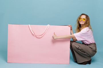 Smiling shopper with giant pink bag giving thumbs up on blue studio background