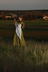 A beautiful woman with long red hair in a vintage green dress on a hill above a field and a village with a candlestick and candles in her hands in the evening