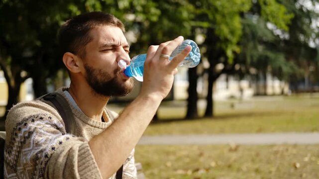 A man with a backpack sits on a park bench in autumn, drinking water and taking a short rest during his walk