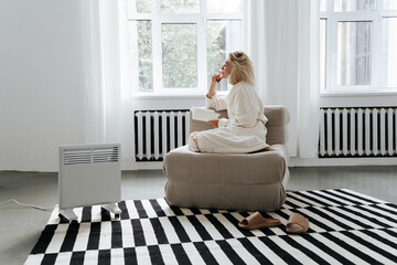 Young woman reading book near electric heater at home