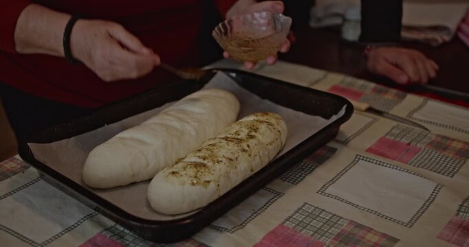 An elderly woman prepares bread with her son. They spread a mixture of olive oil and spices on two loaves of bread. Love, care, and family tradition.