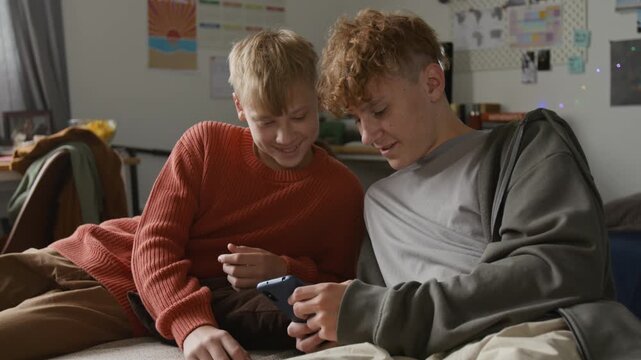 Medium shot of two young teenage boys as brothers using smartphone and watching videos online together smiling while sitting on bed at home