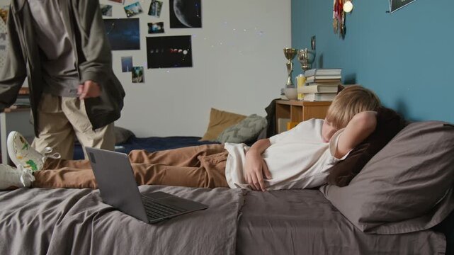 Side view shot of young teenage boy sleeping on bed with laptop with older brother coming up and covering him with blanket, caring family moment